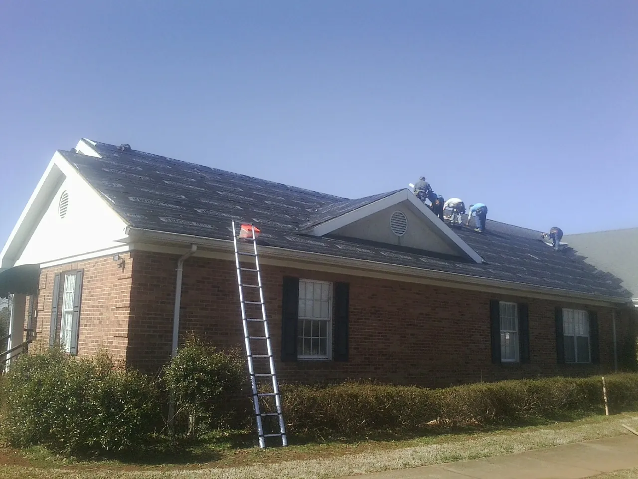 Crew tearing shingles off a brick ranch roof with a ladder set — Emergency Roof Repair work by our crew in Bussey