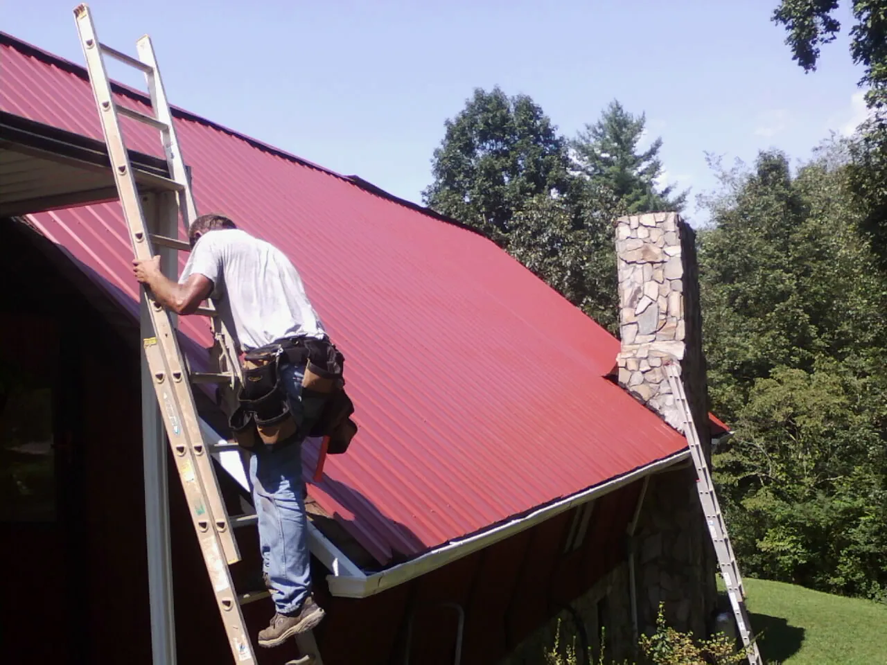 Roofer on a ladder beside a red metal roof and stone chimney — Soffit & Fascia Repair by Roofing Brothers in Bussey